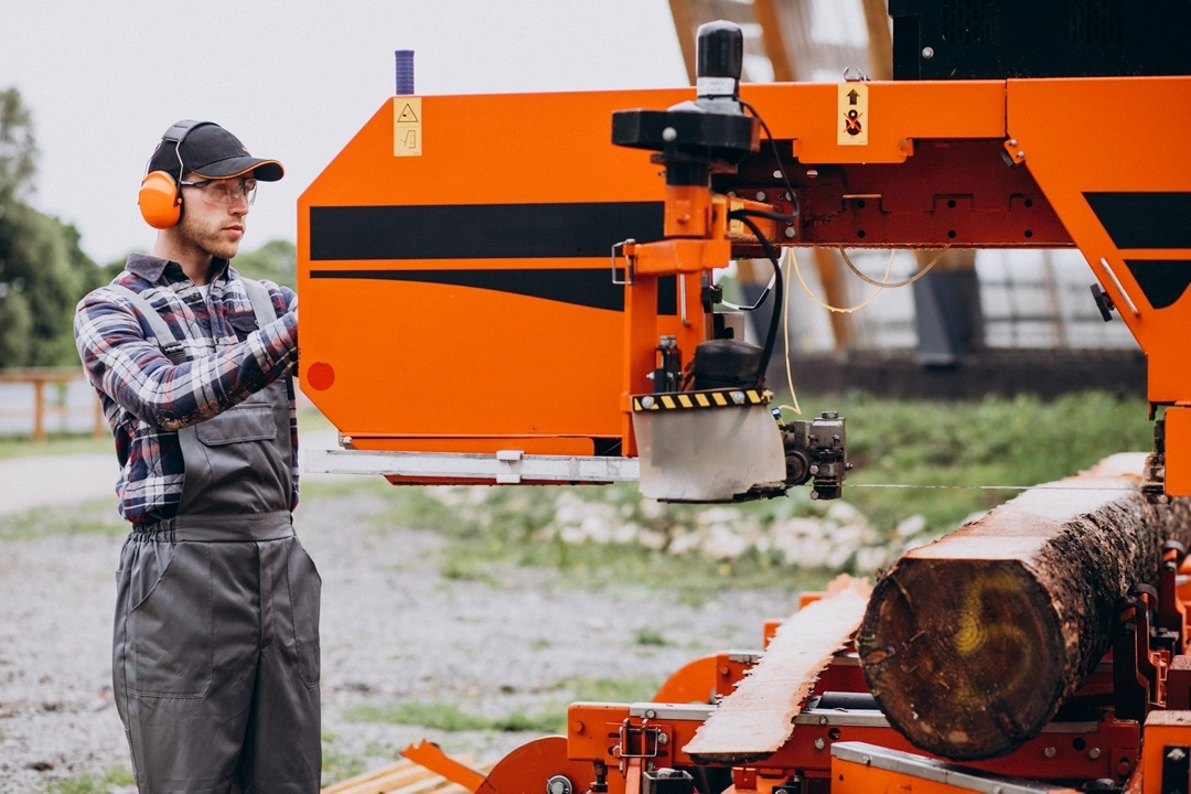 Man wearing protective gear operating an orange portable sawmill machine while cutting a log outdoors