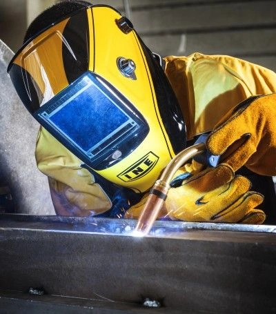 Welder with yellow INE helmet performing MIG welding on metal surface.